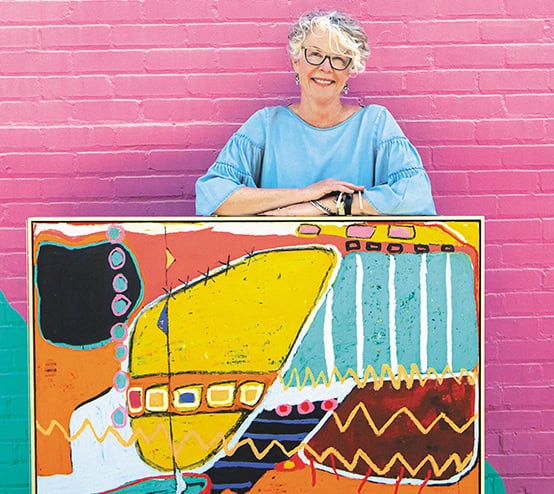 Cindy Walton smiling and standing in front of a pink wall at Pink Dog Creative in Asheville's River Arts District to show off her large, colorful, abstract painting.