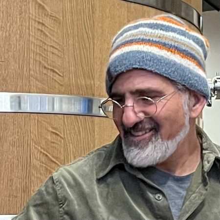 Asheville Artist, Jacob Krueger smiling and looking to the side, wearing a striped beanie, eyeglasses, and a green shirt.