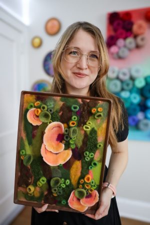 Amy Reader in her studio holding a felted green and orange artpiece featuring natural textures ad chicken of the woods fungi.